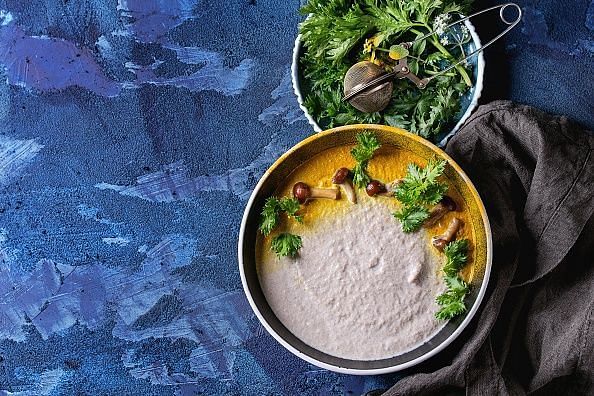Mushroom cream soup in black ceramic bowl served with turmeric powder, forest mushrooms, greens on textile napkin over dark blue texture concrete background. Top view with copy space