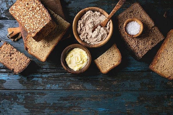 Variety loaves of sliced homemade rye bread whole grain and seeds for breakfast with olive wood bowls of butter, salt, liver paste over old dark wooden background. Top view, copy space.