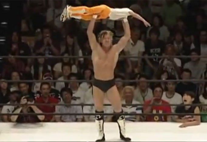 Kenny Omega lifts nine-year-old Karuda in a military press slam.