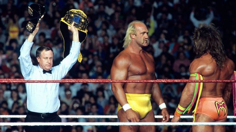 Earl Hebner hefts the WWE world championship overhead as Hulk Hogan and the Ultimate Warrior face off.
