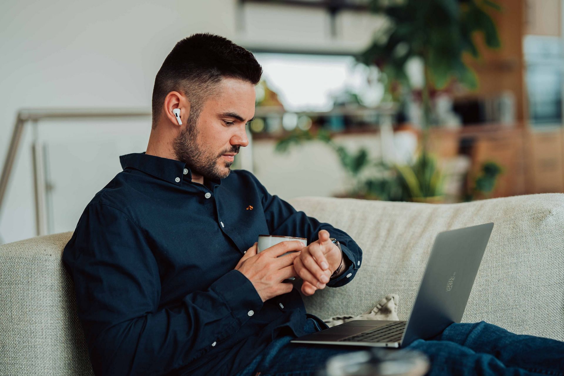 A man sitting on a couch and working on his laptop while drinking a cup of coffee and looking at the time on his watcg