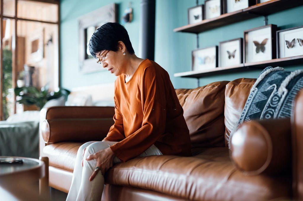 A distraught senior Asian woman feeling unwell, suffering from chronic pain while sitting on sofa in the living room at home