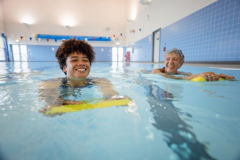 A senior women swimming while using a float alongside a swimming instructor in a swimming pool (Image via Getty Images)