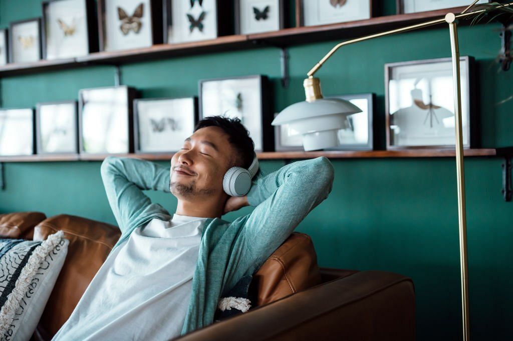 Young Asian man with hands behind head, relaxing on sofa and listening to music with headphones at home