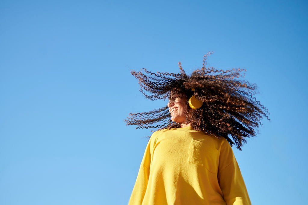 Woman moving her curly hair with blue sky in the background (Image via Getty Images)