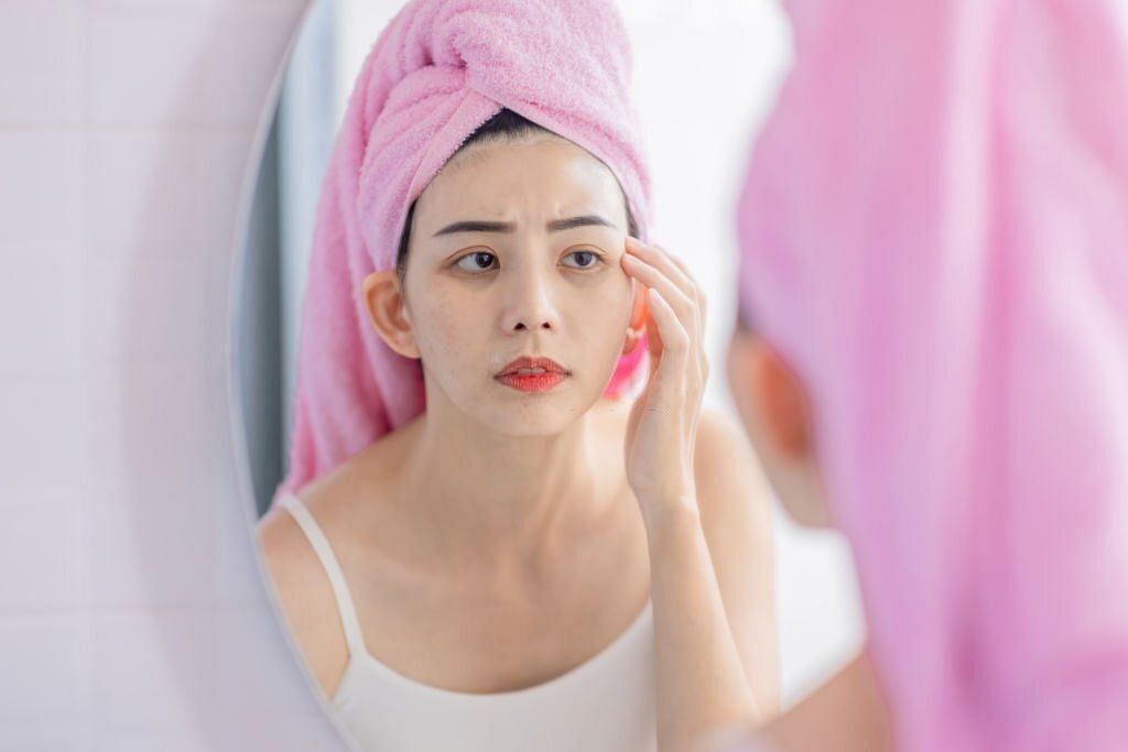 asian woman with towel on head looking mirror feel upset about dark rings under her eyes and touch her face