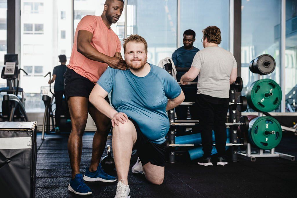 Male fitness instructor assisting overweight man in exercise at gym (Image via Getty Images)