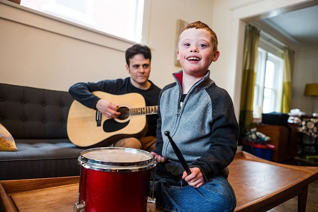 A cute little boy with Down Syndrome playing a drum while his dad plays guitar at home in their living room (authentic). Music therapy is often used with Down Syndrome children(Image via Getty Images)