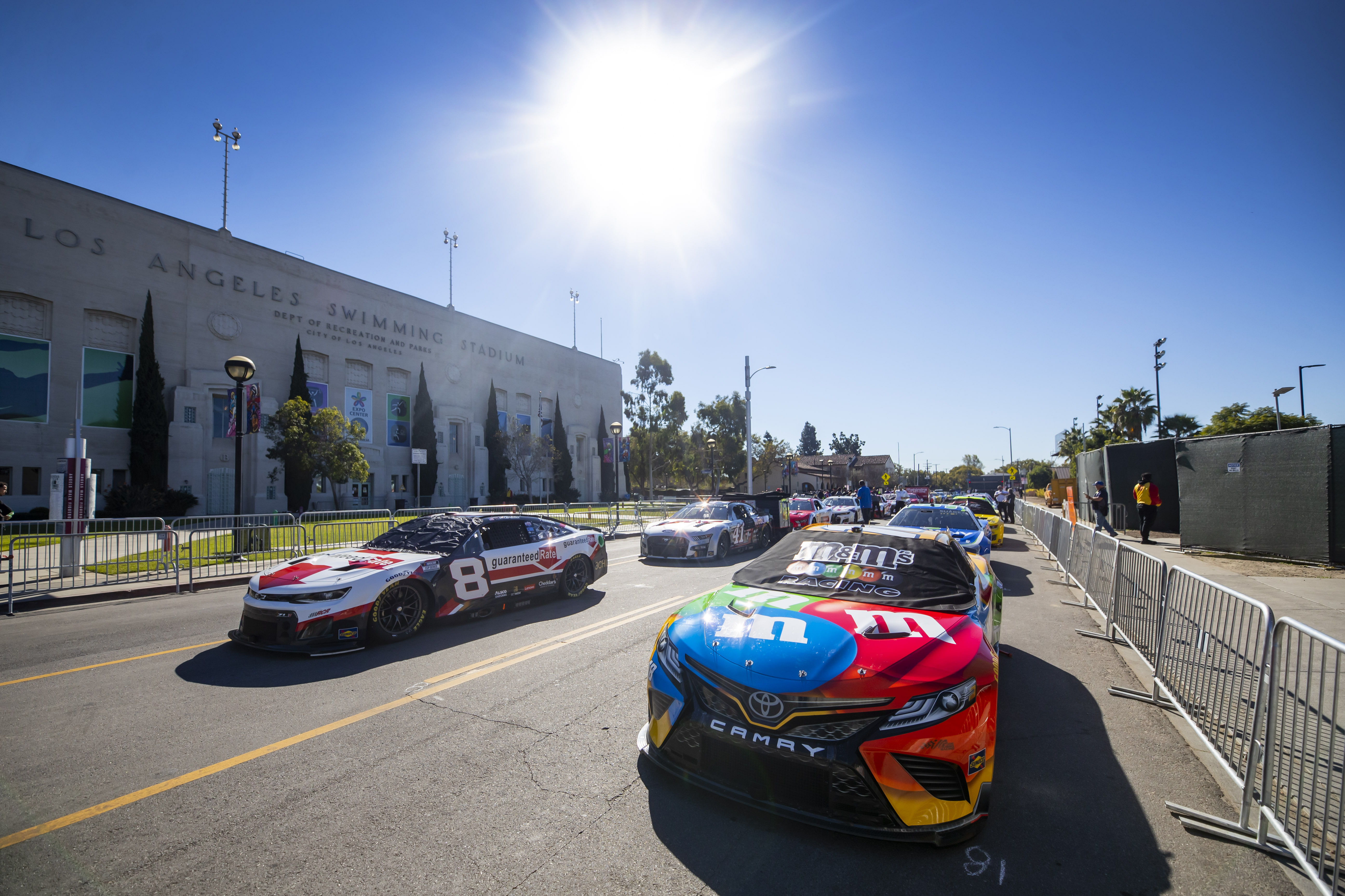 NASCAR Cup Series cars of Kyle Busch (18) and Tyler Reddick (8) sit lined up on Bill Robertson Blvd in front of the Los Angeles Swimming Stadium prior to entering Los Angeles Memorial Coliseum for the Busch Light Clash at The Coliseum. Mandatory Credit: Mark J. Rebilas-Imagn Images