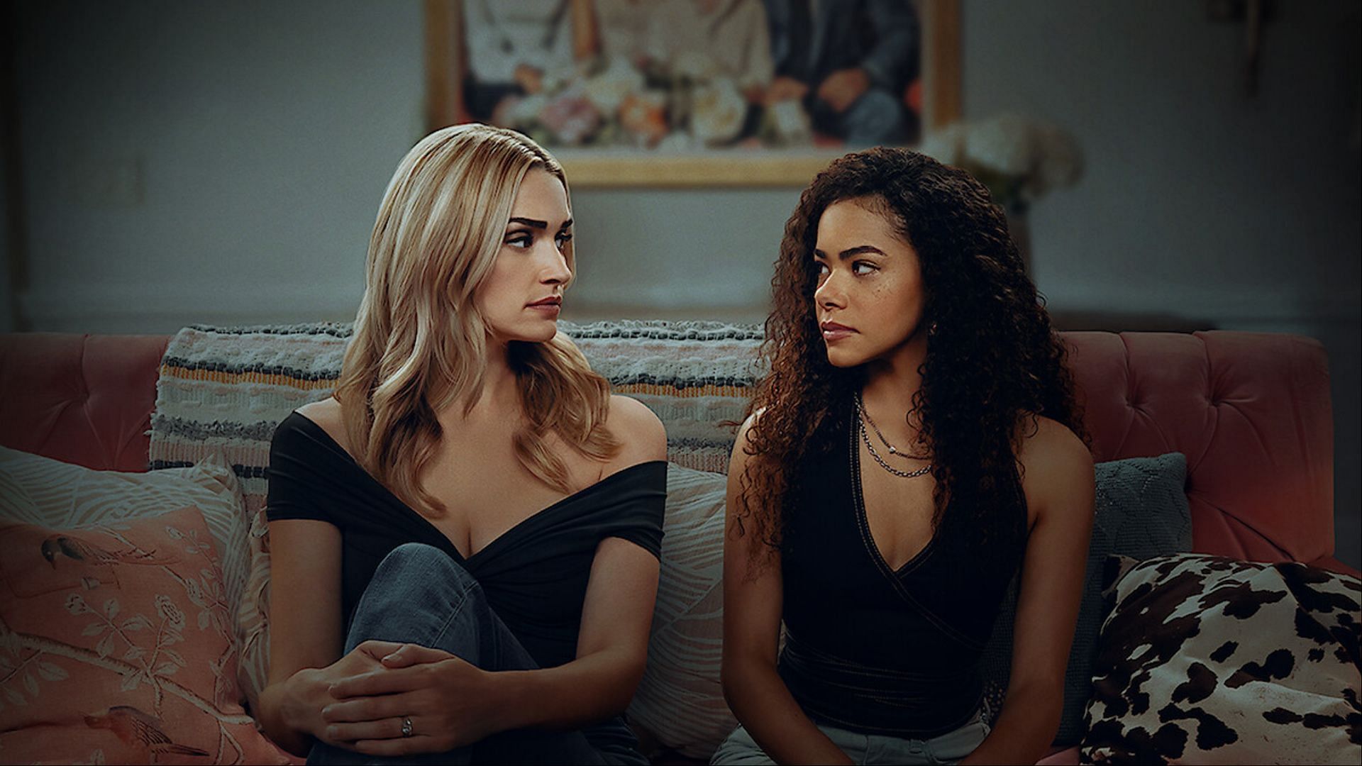 Georgia and Ginny sit side by side on a pink couch, staring at each other with serious expressions in Ginny &amp; Georgia season 3.