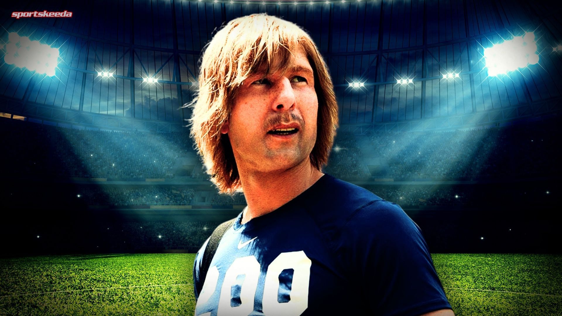 Glen Powell in character as Chad Powers, wearing a football jersey with long hair and a mustache, standing in a stadium under bright lights.