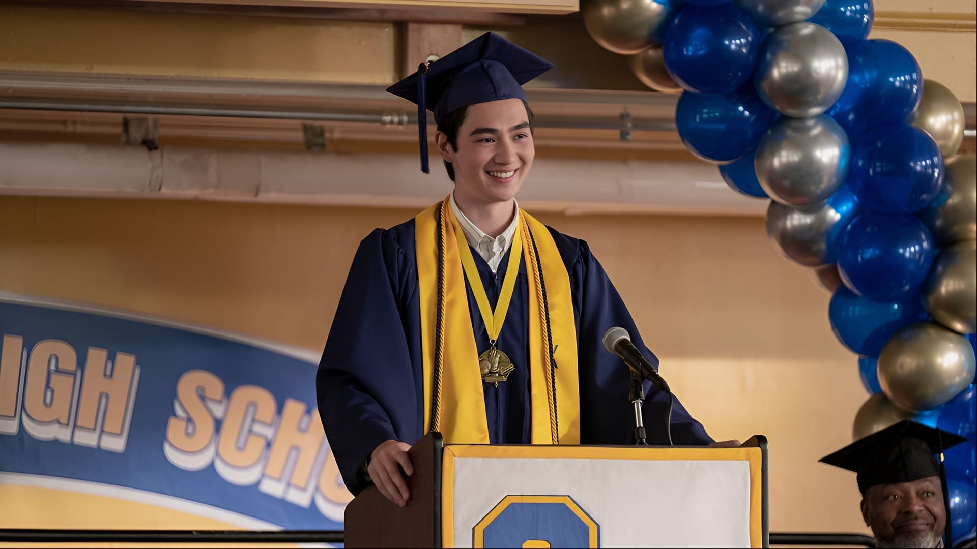 Steven delivers his graduation speech in The Summer I Turned Pretty season 3. Alt text: Steven Conklin smiles at a podium in his graduation cap and gown, giving a speech in The Summer I Turned Pretty. (Image via Amazon Prime)