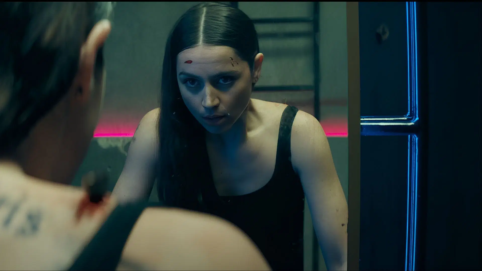 Ana de Armas with blood on her face looking into a mirror inside a neon-lit room.