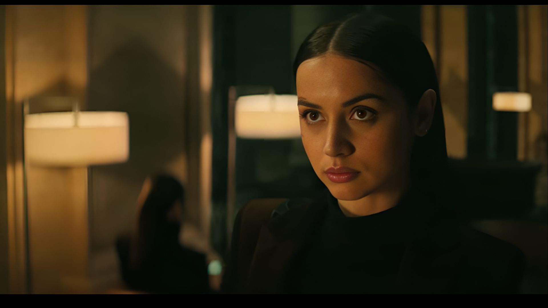 Ana de Armas as Eve Macarro standing in a warmly lit room with a serious expression, two lamps and a mirror visible behind her.
