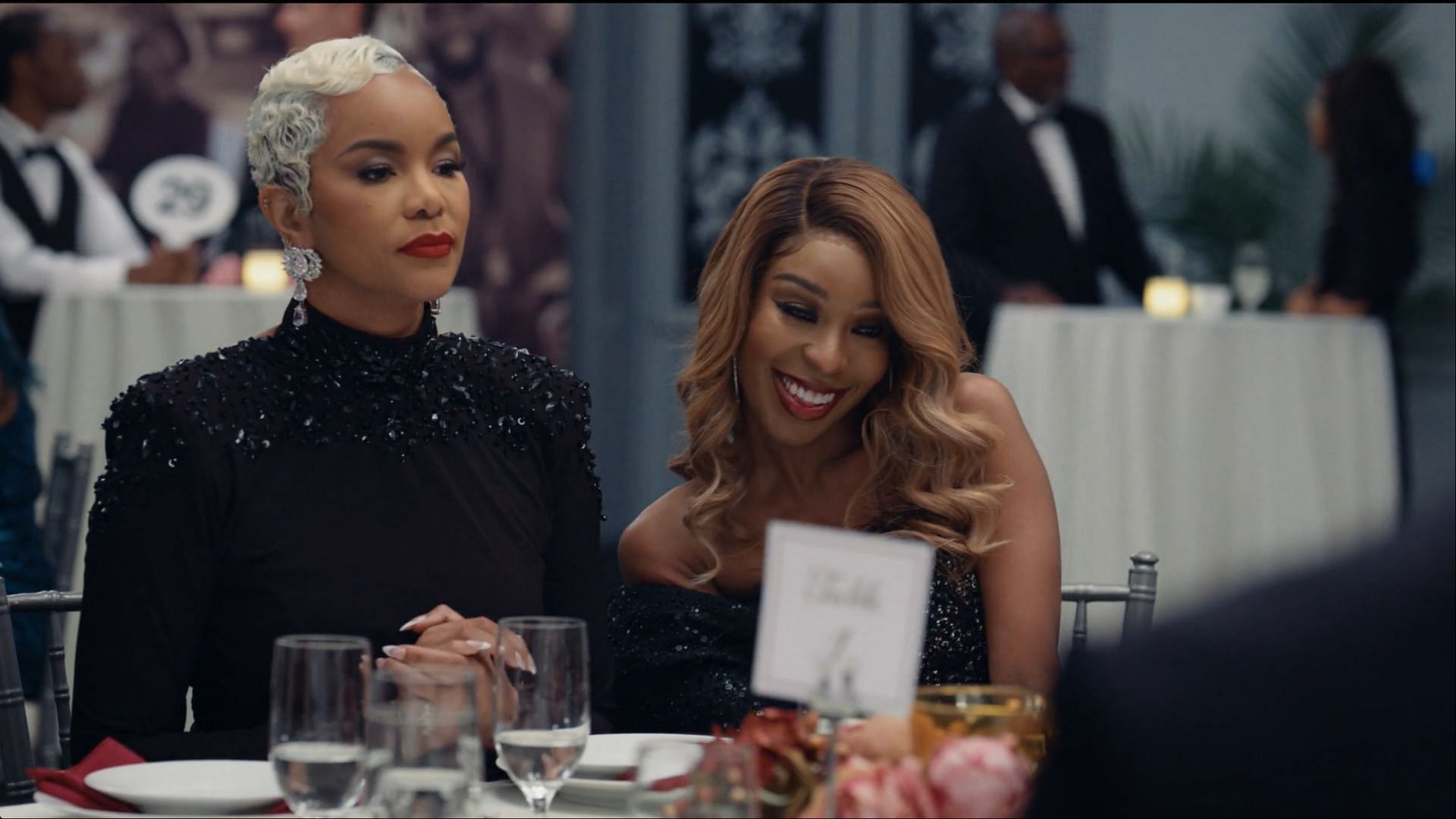 Rasheda and Naomi seated at a banquet table, with Rasheda appearing tense and Naomi smiling warmly.