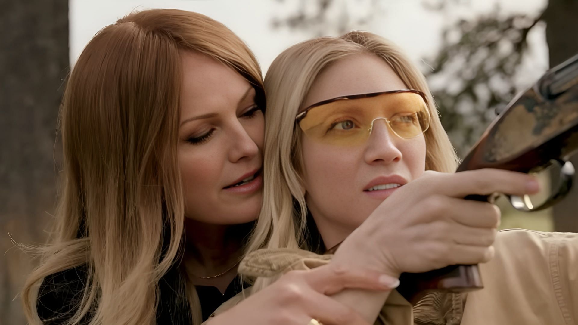 Malin &Aring;kerman as Margo guiding Brittany Snow as Sophie in a shooting lesson scene from The Hunting Wives season 1, both wearing protective glasses outdoors.