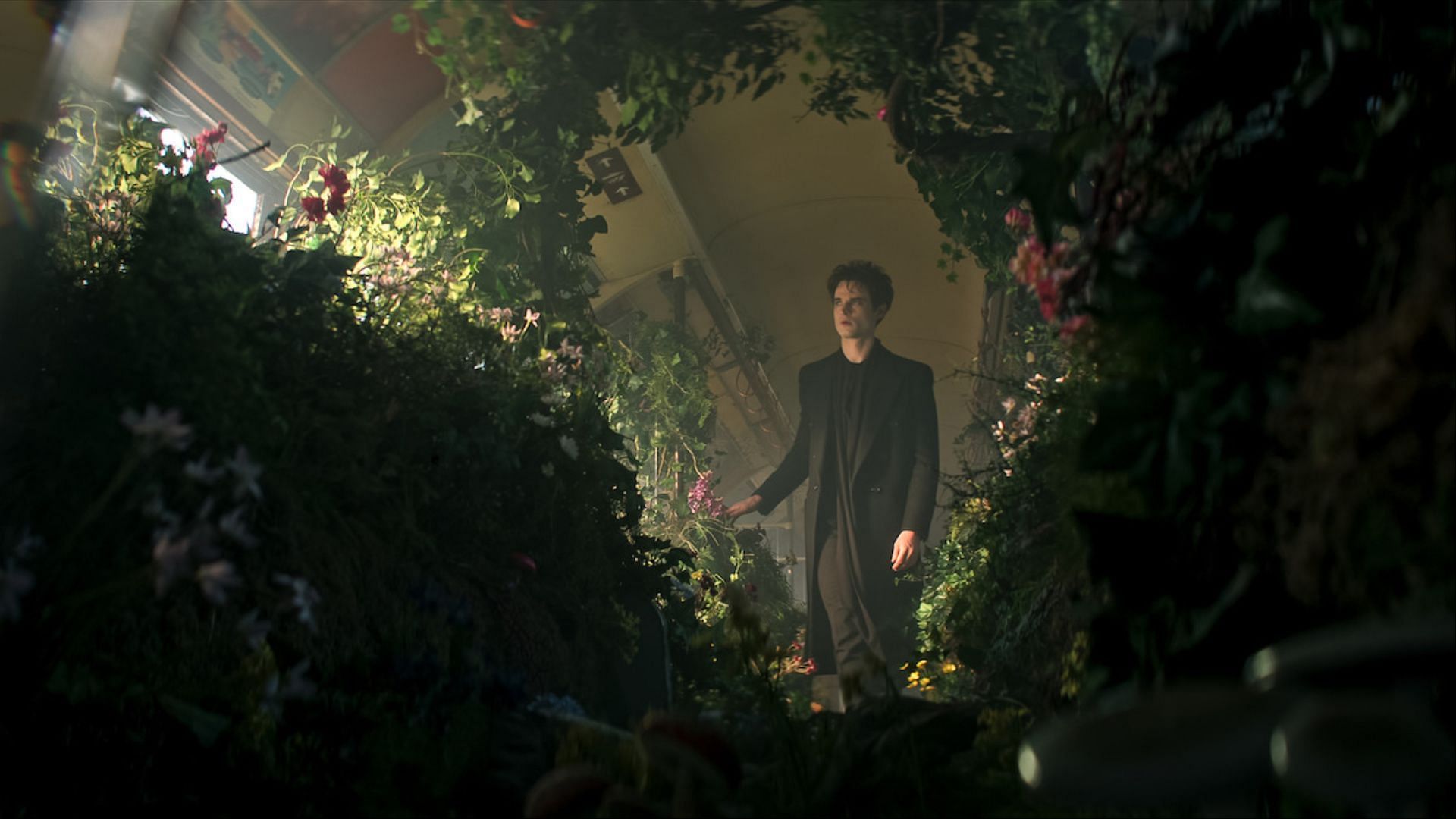 Tom Sturridge as Dream stands in a lush corridor overgrown with plants and flowers in The Sandman season 2, representing the Dreaming.