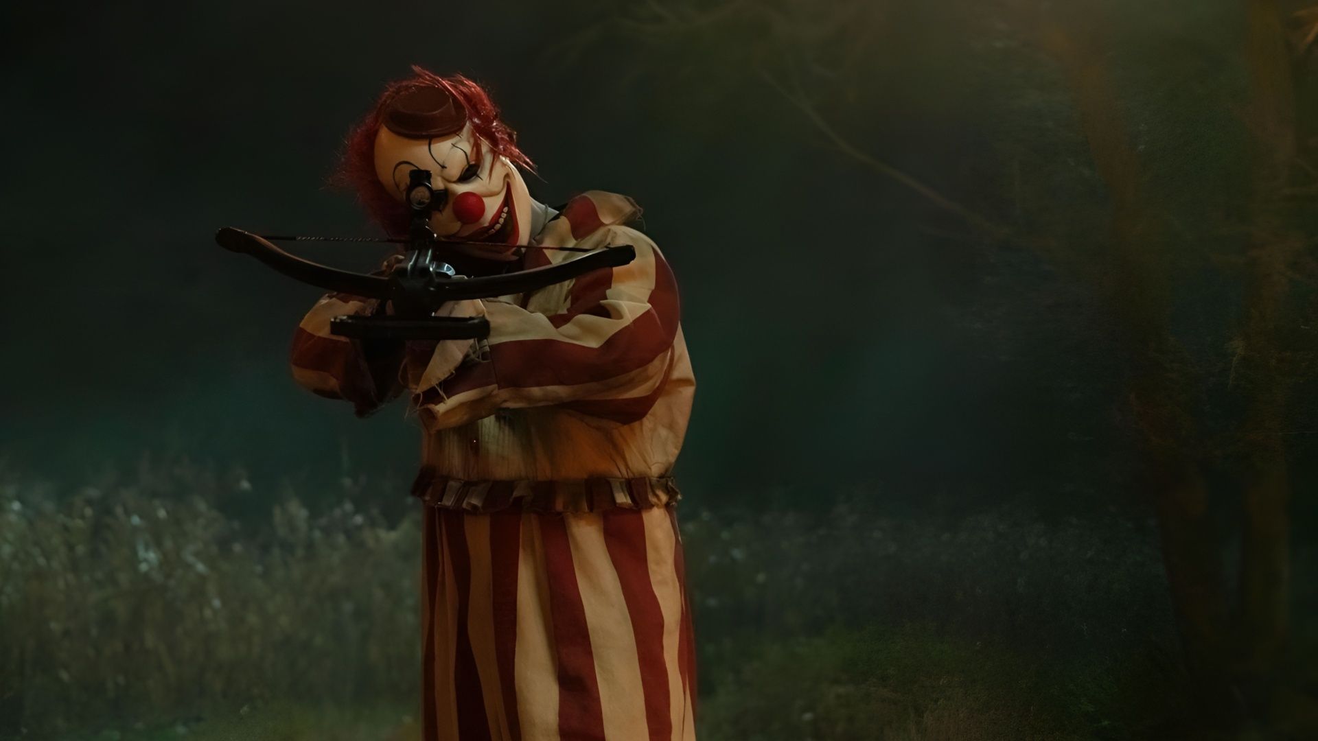 A masked figure dressed as Frendo the Clown aiming a crossbow in a nighttime forest scene from Clown in a Cornfield.