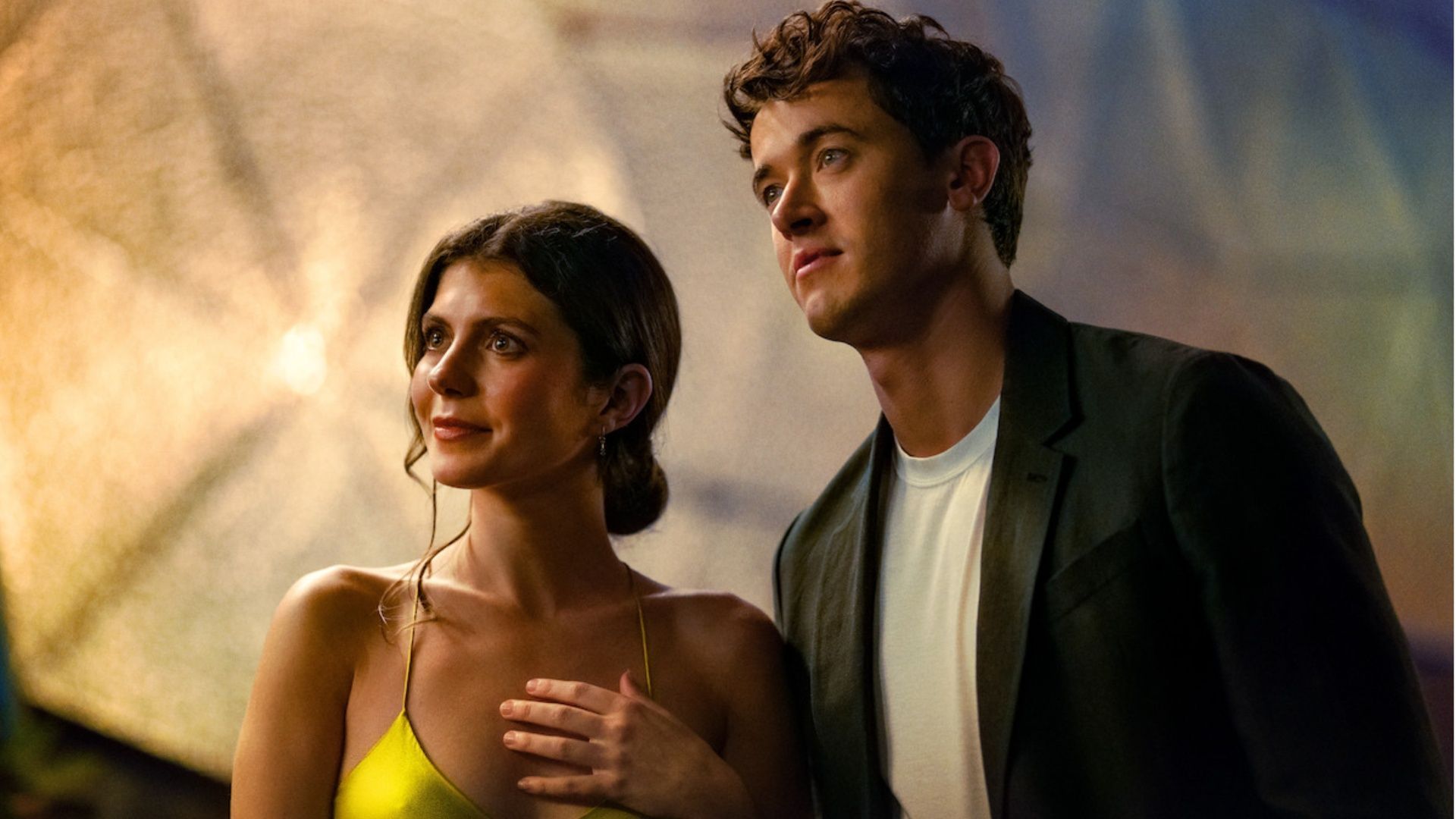 Emily Bader in a yellow dress and Tom Blyth in a dark jacket standing close together at night in a scene from People We Meet on Vacation