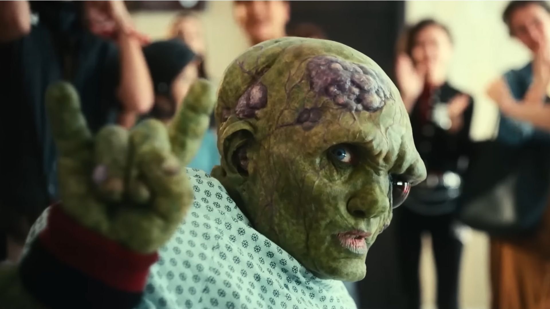 The Toxic Avenger raising his hand in a rock gesture while people clap in the background.