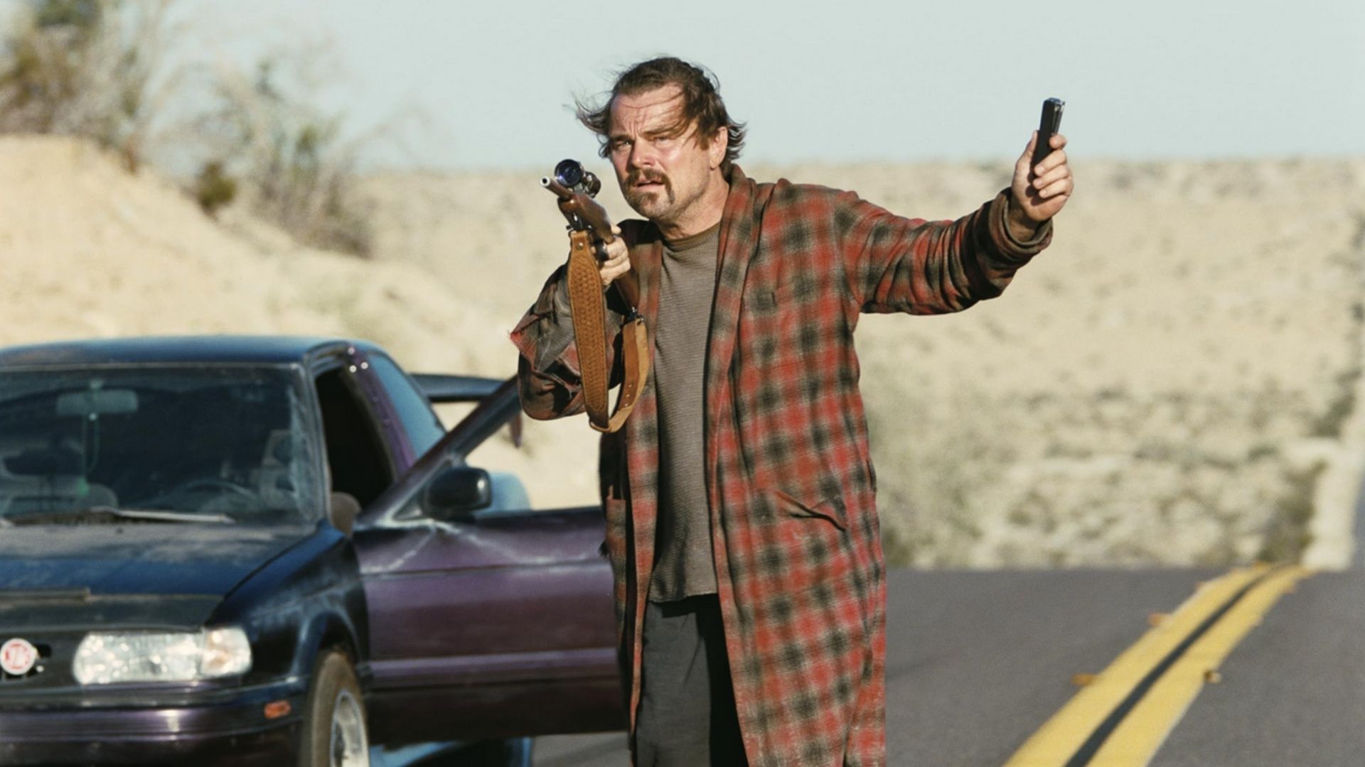 Leonardo DiCaprio in a plaid robe holding a rifle and pistol near a car on a desert road in One Battle After Another.