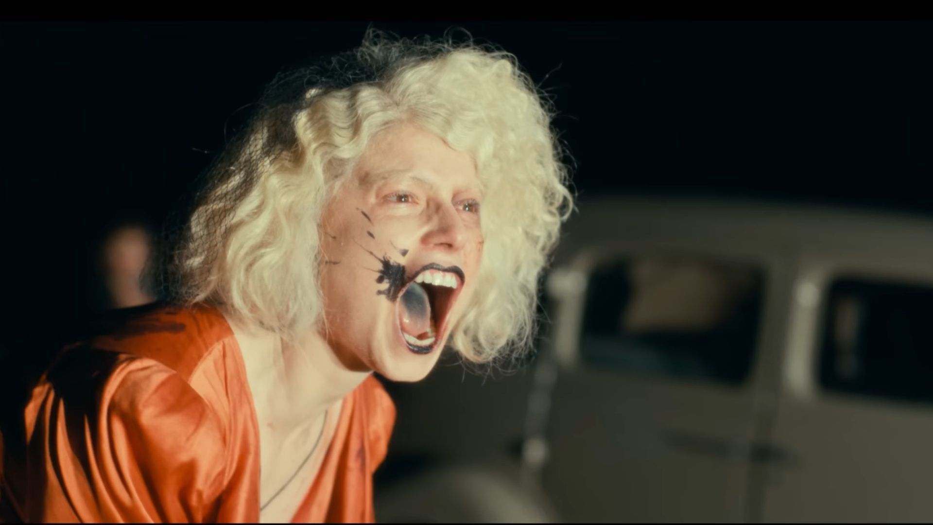 Jessie Buckley as the bride screams with her mouth wide open in a night scene, wearing a red dress, in The Bride trailer.