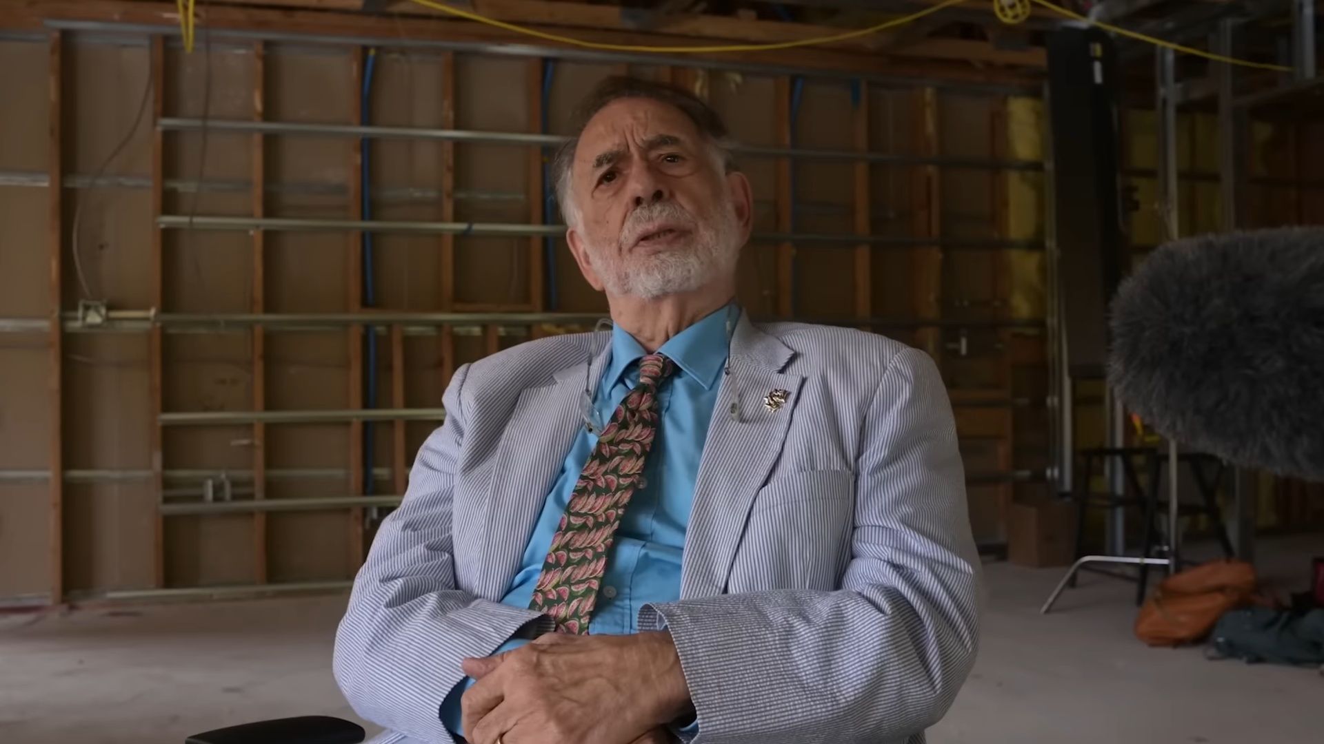 Francis Ford Coppola in a light suit and patterned tie sits in a partially built set while filmed for Megadoc.