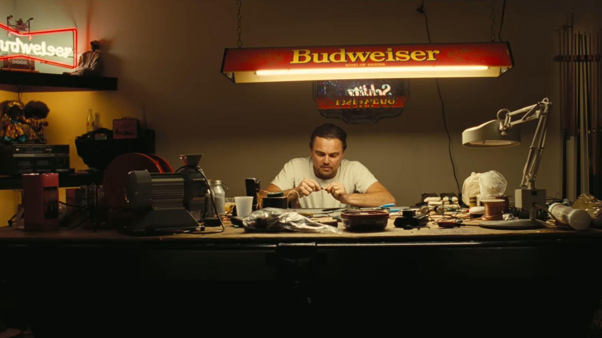 Leonardo DiCaprio sitting under a Budweiser sign while handling tools and wires in One Battle After Another
