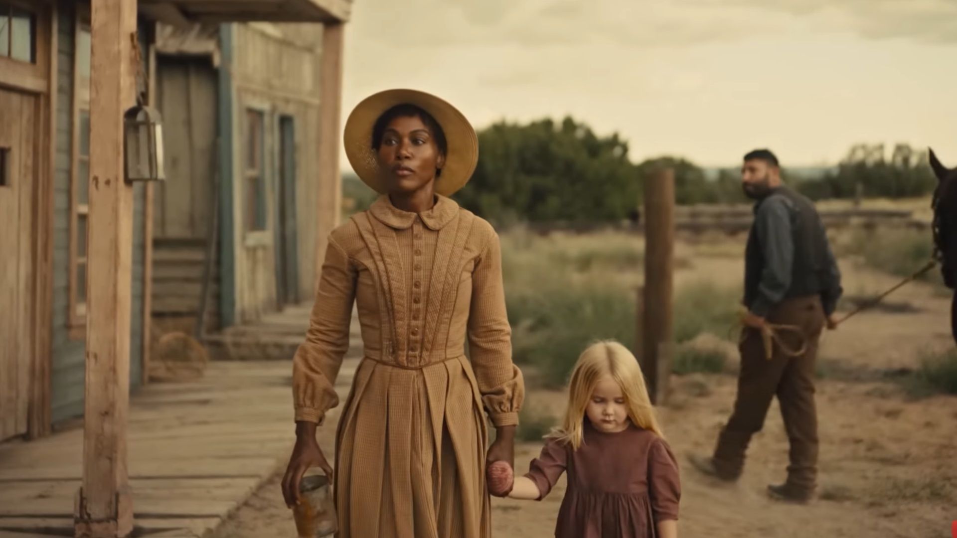 A woman in a brown dress walks hand in hand with her daughter through an old Western town.