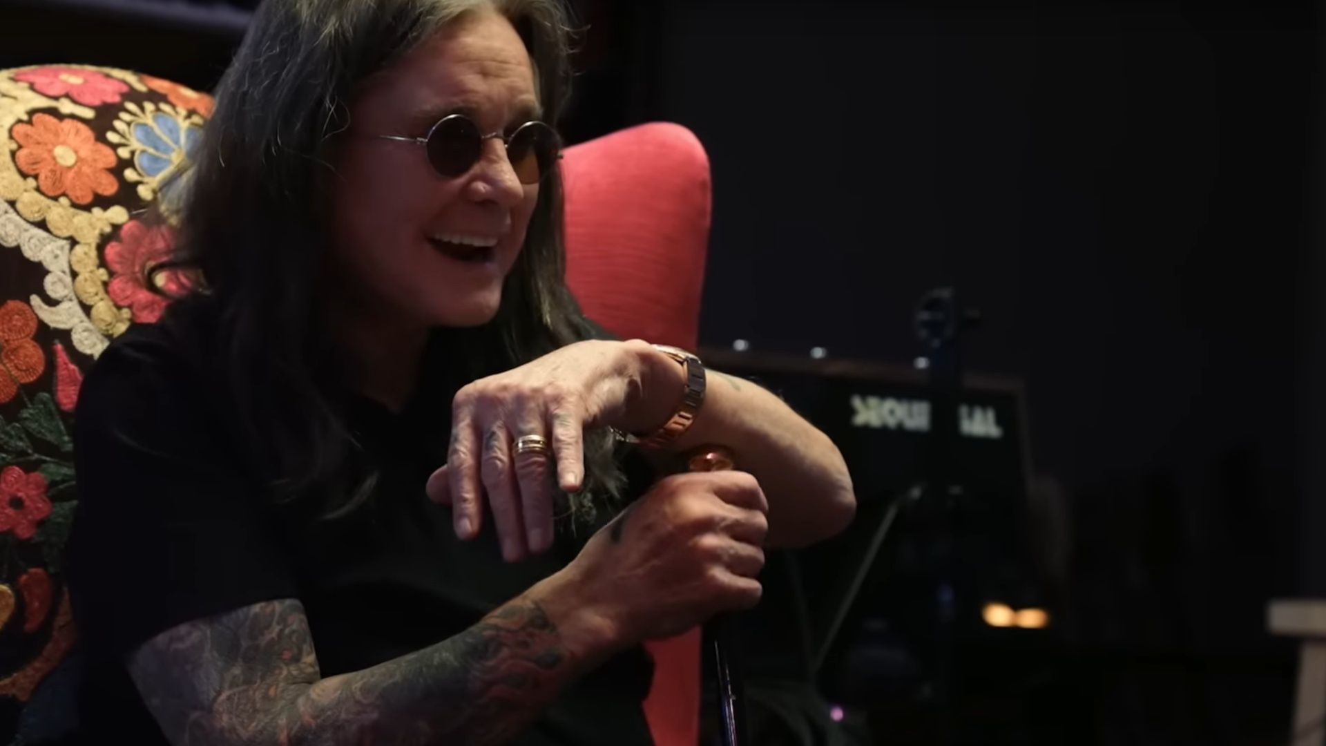 Ozzy Osbourne smiling and seated with a cane, wearing sunglasses, during a recording break in the documentary.