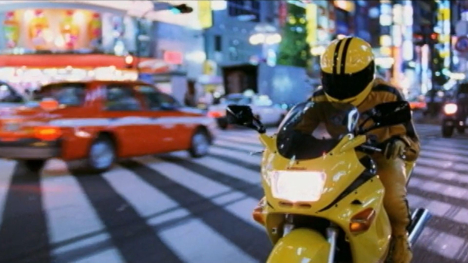 The Bride rides a yellow motorcycle through a city at night. (Image via A Band Apart)