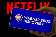 Warner Bros. Discovery Logo Displayed on Smartphone Against Netflix Background (Image via Getty)