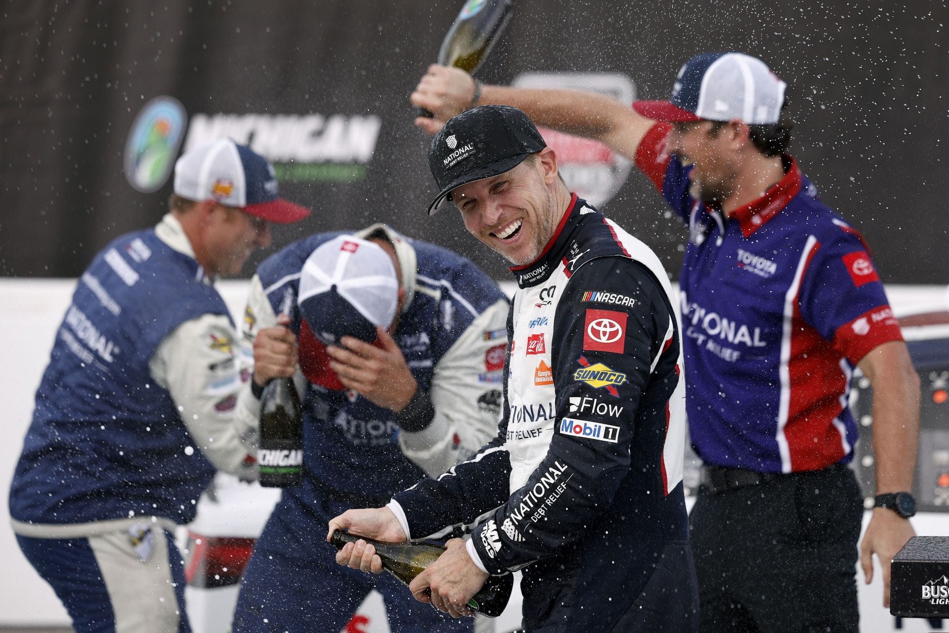 Denny Hamlin, driver of the #11 National Debt Relief Toyota, and crew spray champagne in victory lane after winning the NASCAR Cup Series FireKeepers Casino 400 at Michigan International Speedway on June 07, 2026 in Brooklyn, Michigan. - Source: Getty